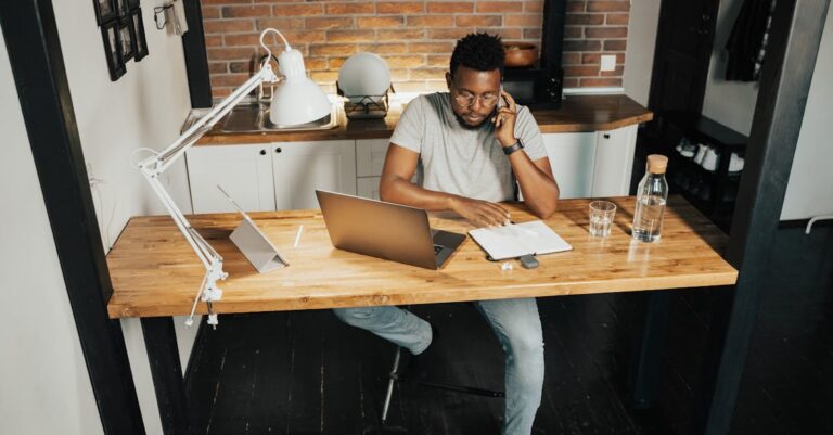 A young man at a desk in a home office, using a laptop and phone, embodying remote work lifestyle.