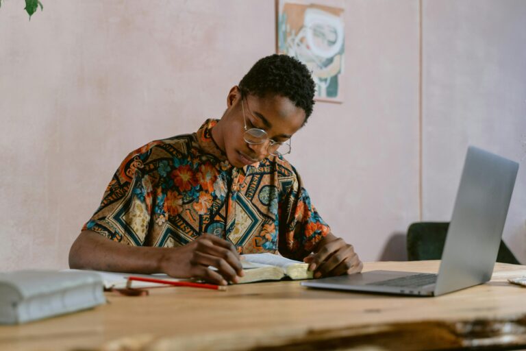 Young adult studying with a book and laptop indoors, wearing colorful shirt.