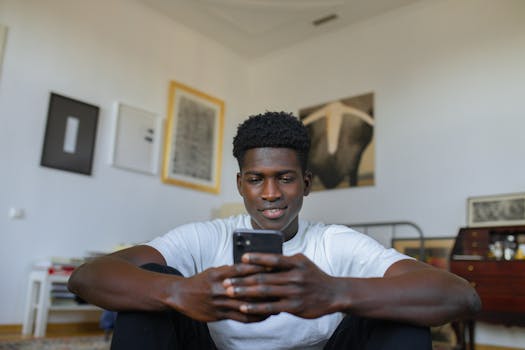 Young man in a white shirt is sitting indoors, smiling while using a smartphone.